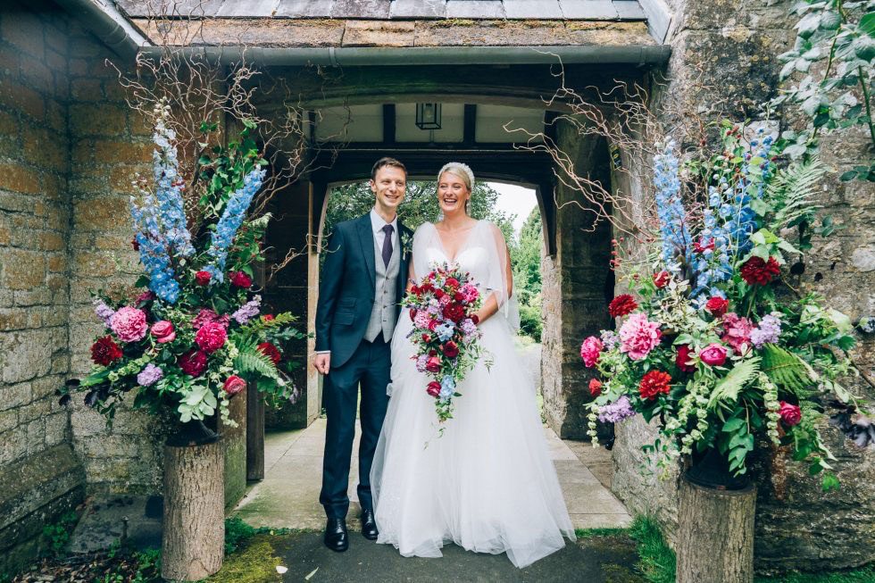 Bride and groom in a stone porch flanked by two tall arrangements of blue delphiniums, pink peonies and red roses on log plinths