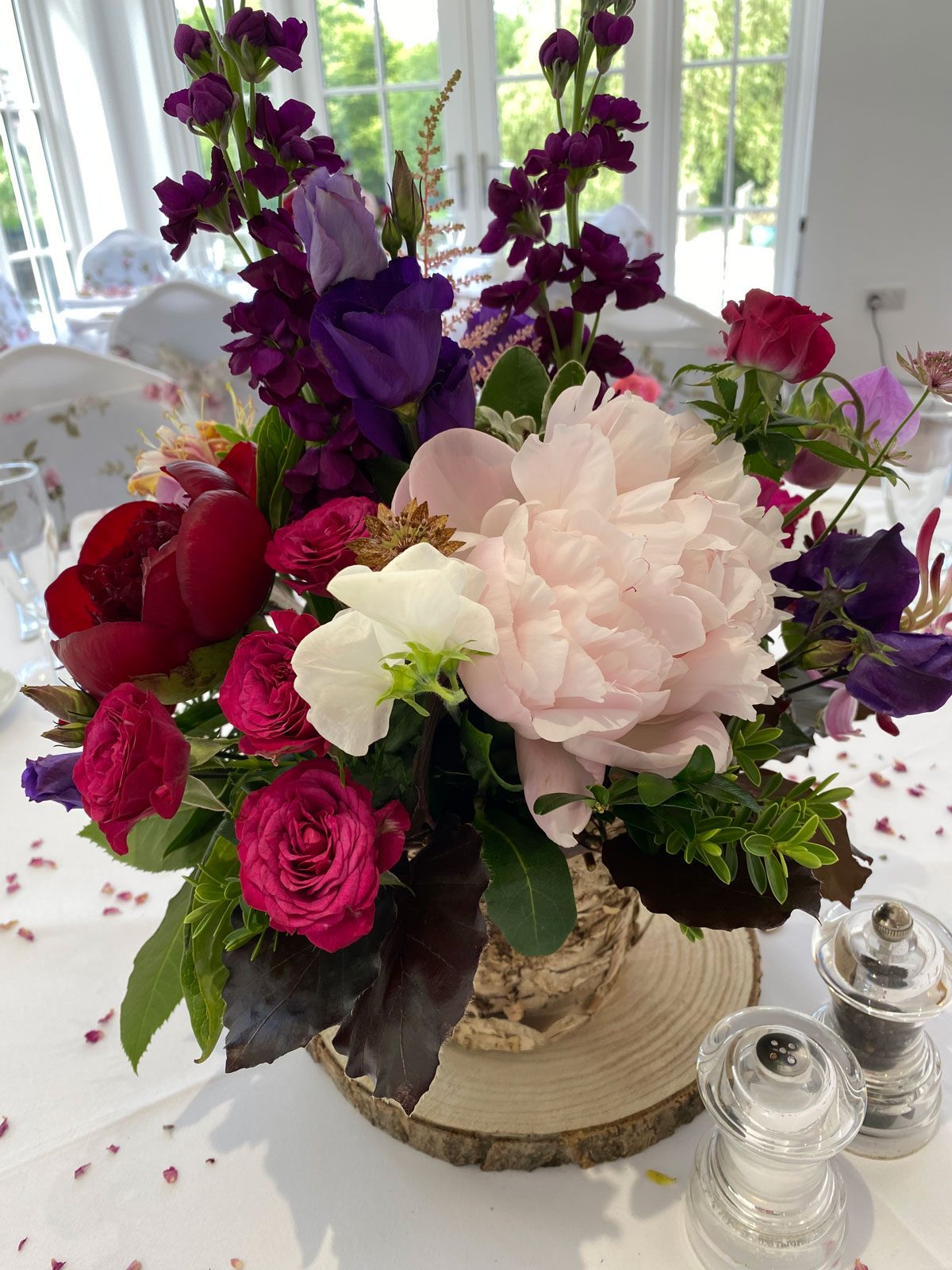 Table centrepiece of blush peonies, deep pink roses and plum stocks on a log slice